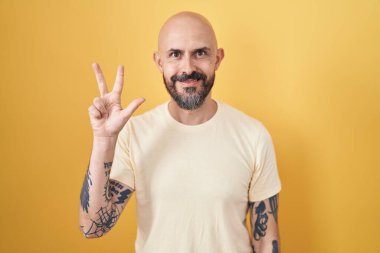 Hispanic man with tattoos standing over yellow background showing and pointing up with fingers number three while smiling confident and happy. 