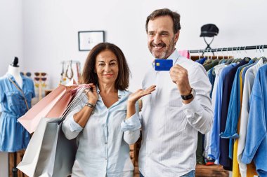 Hispanic middle age couple holding shopping bags and credit card smiling cheerful presenting and pointing with palm of hand looking at the camera. 