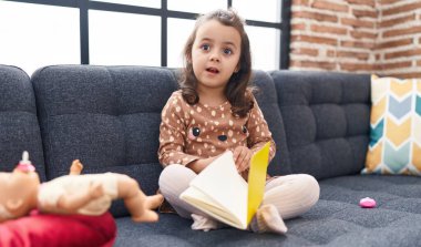 Adorable hispanic girl reading book sitting on sofa at home