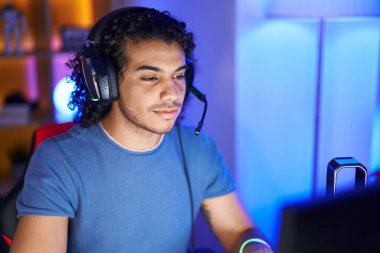Young latin man streamer sitting on table with serious expression at gaming room