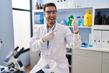 Young hispanic man with beard working at scientist laboratory holding green ribbon smiling happy pointing with hand and finger 