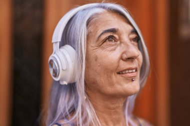 Orta yaşlı, kır saçlı, gülümseyen, kendine güvenen, sokakta müzik dinleyen bir kadın.