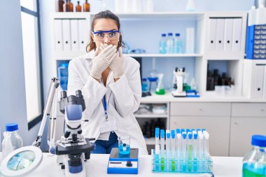 Young hispanic woman working at scientist laboratory shocked covering mouth with hands for mistake. secret concept. 