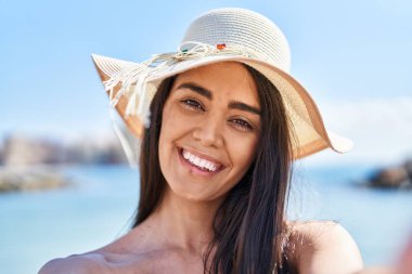 Genç İspanyol kadın, deniz kenarında kendine güvenen bikini ve yaz şapkası takarak gülümsüyor.