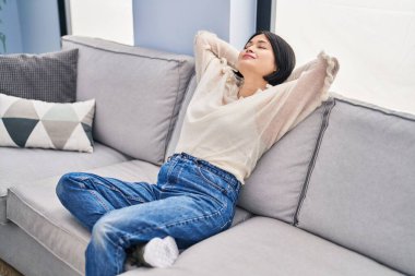 Young chinese woman relaxed with hands on head sitting on sofa at home
