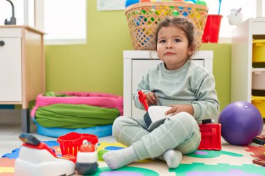 Adorable hispanic toddler playing with supermarket toy sitting on floor at kindergarten