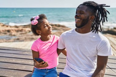 Father and daughter using smartphone hugging each other at seaside
