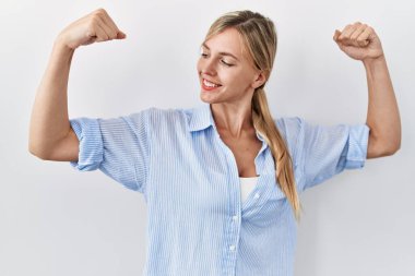 Beautiful blonde woman standing over white background showing arms muscles smiling proud. fitness concept. 