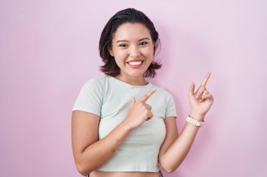 Hispanic young woman standing over pink background smiling and looking at the camera pointing with two hands and fingers to the side. 