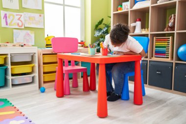 Adorable hispanic boy preschool student sitting on table drawing on paper at kindergarten