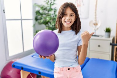 Young hispanic girl holding small rehabilitation ball pointing thumb up to the side smiling happy with open mouth 