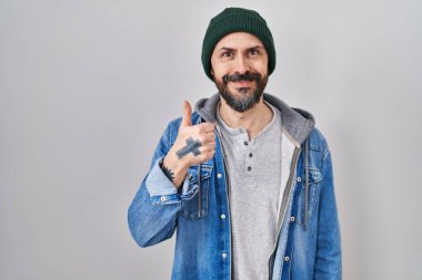 Young hispanic man with tattoos wearing wool cap doing happy thumbs up gesture with hand. approving expression looking at the camera showing success. 