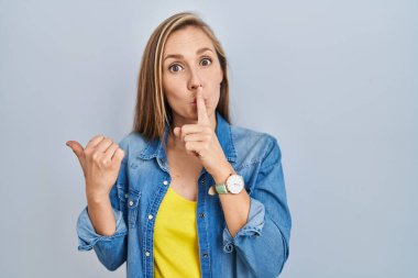 Mavi arka planda dikilen genç sarışın kadın sessiz olmak istiyor. Parmakları dudaklarında, elleri yandan işaret ederek. sessizlik ve gizli konsept. 