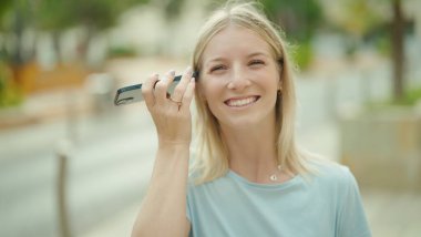 Young blonde woman smiling confident listening audio message by the smartphone at street