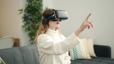 Evde sanal gerçeklik gözlüğü kullanarak video oyunu oynayan genç sarışın kadın.