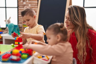 Teacher with boy and girl playing with construction blocks sitting on table at kindergarten