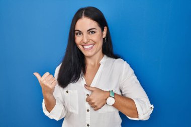 Young hispanic woman standing over blue background pointing to the back behind with hand and thumbs up, smiling confident 