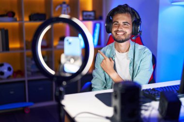 Young hispanic man with tattoos recording tutorial with smartphone smiling cheerful pointing with hand and finger up to the side 