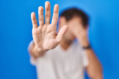 Mavi arka planda duran İspanyol genç adam elleriyle gözlerini kapatıyor ve üzgün ve korku ifadesiyle dur hareketi yapıyor. utangaç ve olumsuz kavram. 