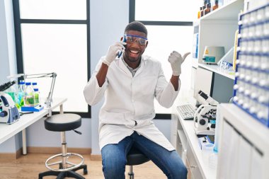 African american man working at scientist laboratory speaking on the phone pointing thumb up to the side smiling happy with open mouth 