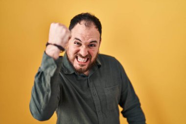 Plus size hispanic man with beard standing over yellow background angry and mad raising fist frustrated and furious while shouting with anger. rage and aggressive concept. 