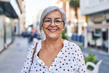 Orta yaşlı bir kadın sokakta kendine güvenerek gülümsüyor.