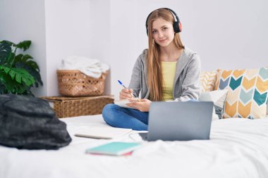 Young caucasian woman writing on notebook listening to music studying at bedroom