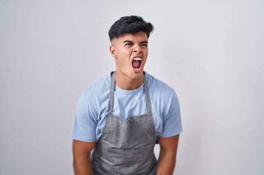 Hispanic young man wearing apron over white background angry and mad screaming frustrated and furious, shouting with anger. rage and aggressive concept. 