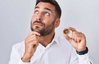 Handsome hispanic man holding cardano cryptocurrency coin serious face thinking about question with hand on chin, thoughtful about confusing idea 