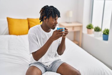 African american man drinking cup of coffee sitting on bed at bedroom