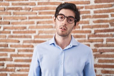 Young hispanic man standing over brick wall background looking sleepy and tired, exhausted for fatigue and hangover, lazy eyes in the morning. 