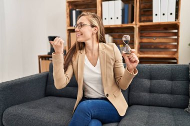 Terapi ofisinde çalışan genç sarışın kadın elinde kum saati tutarak baş parmağını yukarı kaldırıyor ve ağzı açık gülümsüyor. 