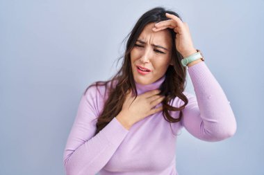 Genç esmer kadın mavi arka planda duruyor. Hastalık ve ateş için alnına dokunuyor. Grip ve soğuk. 