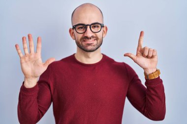 Beyaz arka planda duran sakallı genç kel bir adam gözlük takıyor ve yedi numaralı parmaklarıyla işaret ederken kendinden emin ve mutlu bir şekilde gülümsüyor.. 