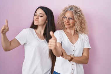 Anne ve kızı pembe arka planda bir arada duruyorlar gururlu görünüyorlar, gülümsüyorlar yan tarafa başparmak işareti yapıyorlar. 