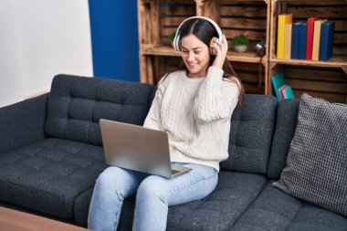 Evdeki kanepede oturan dizüstü bilgisayar ve kulaklık kullanan genç bir kadın.