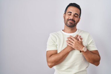 Hispanic man with beard standing over isolated background smiling with hands on chest with closed eyes and grateful gesture on face. health concept. 