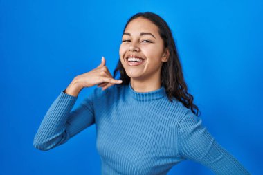 Young brazilian woman standing over blue isolated background smiling doing phone gesture with hand and fingers like talking on the telephone. communicating concepts. 