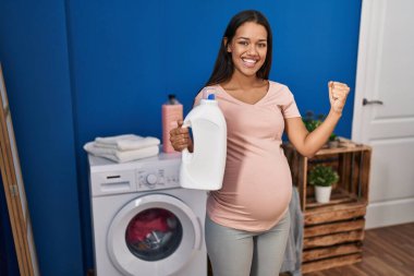 Young pregnant woman doing laundry holding detergent bottle screaming proud, celebrating victory and success very excited with raised arms 