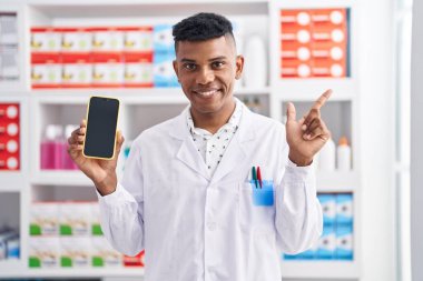 Young hispanic man working at pharmacy drugstore showing smartphone screen smiling happy pointing with hand and finger to the side 