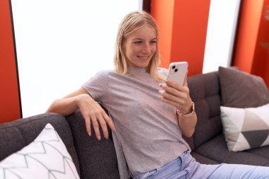 Evdeki kanepede oturan akıllı telefon kullanan genç sarışın kadın.