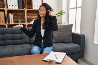 Young asian woman at consultation office confused and annoyed with open palm showing copy space and pointing finger to forehead. think about it. 