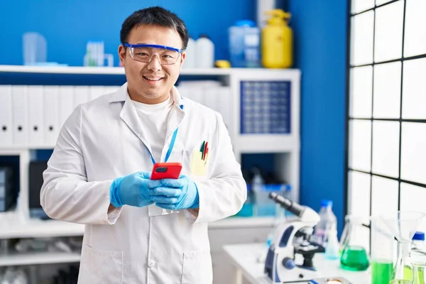 Young chinese man wearing scientist uniform using smartphone at laboratory