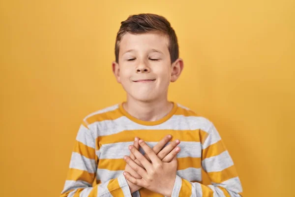 黄色の背景に立っている若い白人の子供は 目を閉じて胸に手を笑顔で 顔に感謝のジェスチャーをしています 健康の概念 — ストック写真