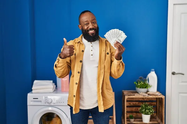 African American Man Holding Money Laundry Room Smiling Happy Positive — Zdjęcie stockowe