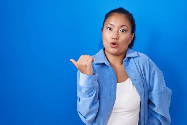 Asian young woman standing over blue background surprised pointing with hand finger to the side, open mouth amazed expression.