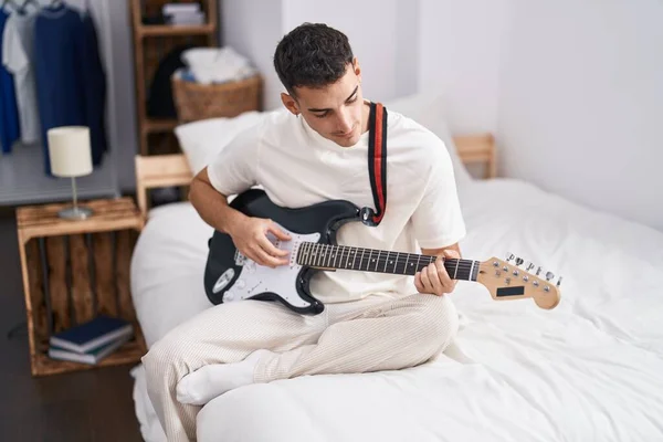Young hispanic man playing electrical guitar sitting on bed at bedroom