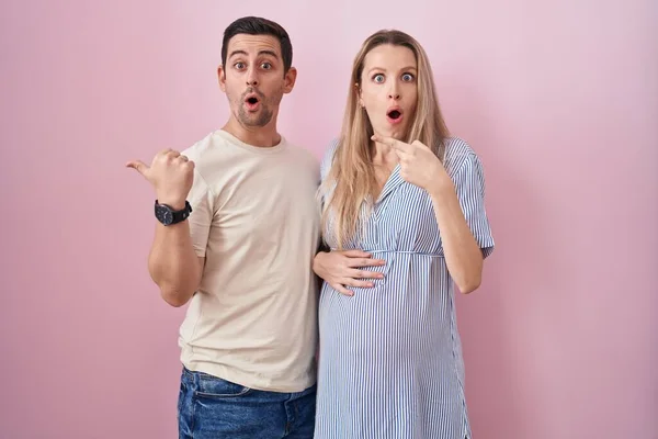Casal Jovem Esperando Bebê Sobre Fundo Rosa Surpreso Apontando Com — Fotografia de Stock