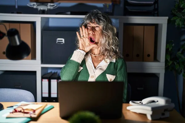 Middle age woman working at night using computer laptop shouting and screaming loud to side with hand on mouth. communication concept.