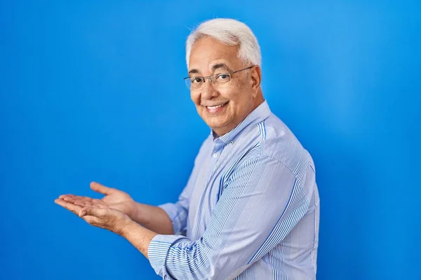 Hispanic senior man wearing glasses pointing aside with hands open palms showing copy space, presenting advertisement smiling excited happy
